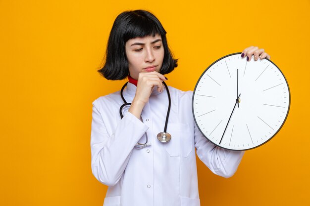 Confused young pretty caucasian woman in doctor uniform with stethoscope holding and looking at clock putting hand on her chin 