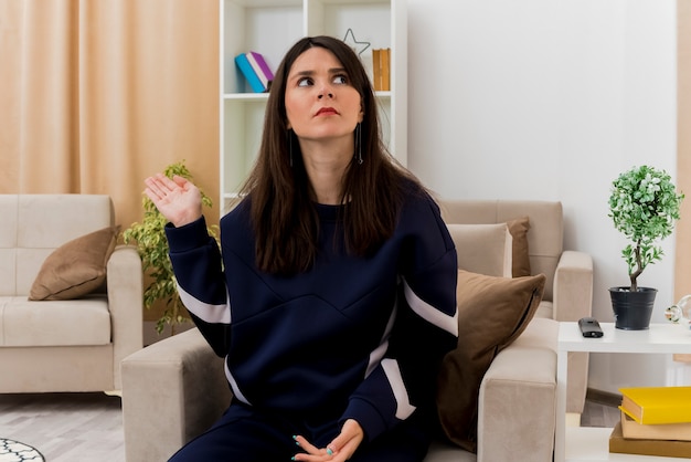 Confused young pretty caucasian woman sitting on armchair in designed living room looking at side pointing with hand at side