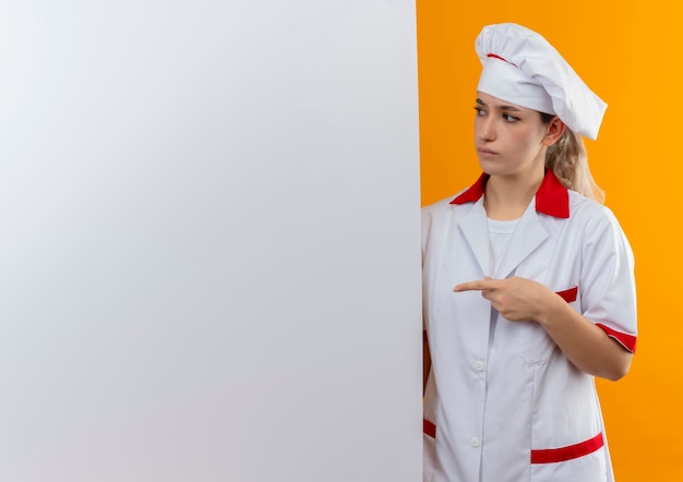 Смущенный молодой симпатичный повар в униформе шеф-повара стоит позади, указывая и глядя на белую стену, изолированную на оранжевой стене с копией пространства
