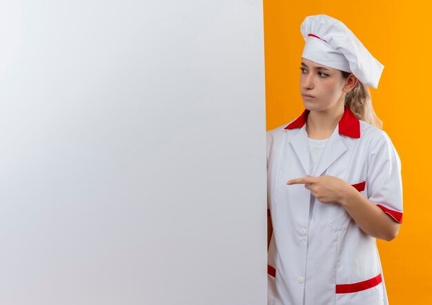 Confused young pretty cook in chef uniform standing behind pointing and looking at white wall isolated on orange wall with copy space