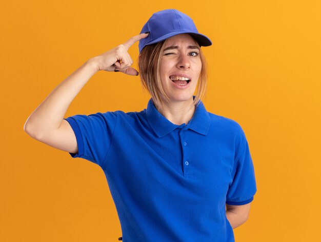 Confused young pretty delivery girl in uniform puts finger on head and looks at camera on orange