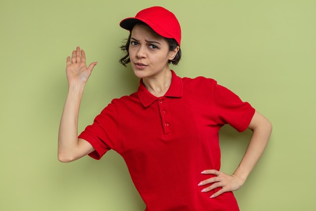 Confused young pretty delivery woman standing with raised hand 