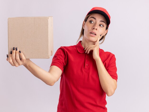 Confused young pretty delivery woman in uniform puts hand on face holding and looking at cardbox isolated on white wall