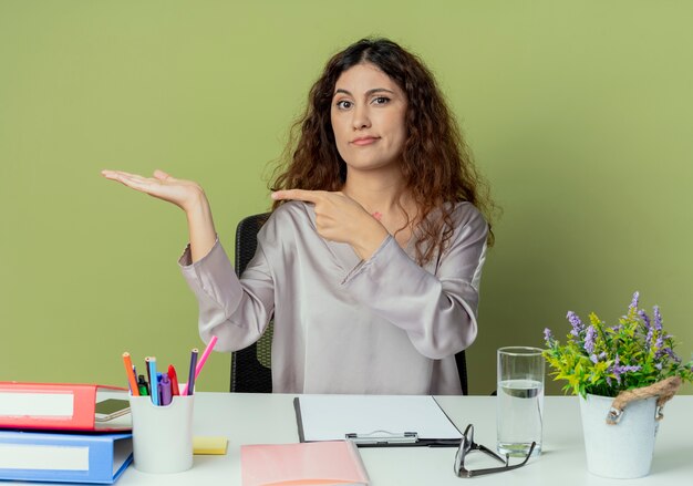 Смущенная молодая симпатичная женщина-офисный работник сидит за столом с офисными инструментами, делая вид, что держит что-то изолированное на оливковом фоне