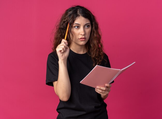 Confused young pretty girl holding pencil and notebook doing think gesture looking at side 