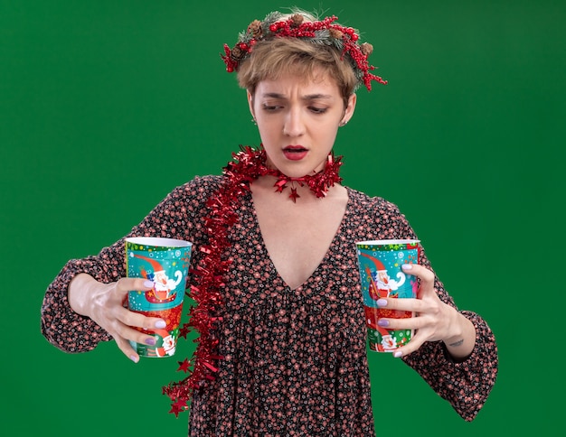 Бесплатное фото Смущенная молодая красивая девушка в рождественском венке и гирлянде из мишуры на шее держит пластиковые рождественские чашки, глядя на одну из них, изолированную на зеленой стене