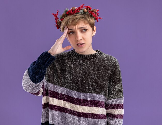 Confused young pretty girl wearing christmas head wreath looking at side touching head isolated on purple wall