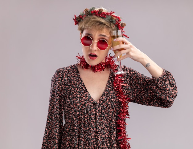 Confused young pretty girl wearing christmas head wreath and tinsel garland around neck with glasses touching head with glass of champagne looking at camera isolated on white background