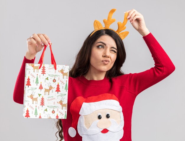 Confused young pretty girl wearing reindeer antlers headband and santa claus sweater holding christmas gift bag grabbing headband looking at side with one eye closed isolated on white wall