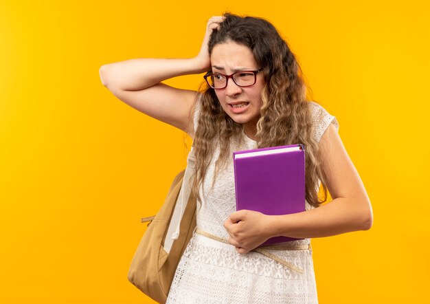 Confused young pretty schoolgirl wearing glasses and back bag holding book looking at side isolated on yellow  with copy space