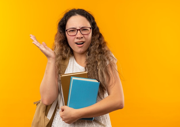 Foto gratuita scolara giovane graziosa confusa con gli occhiali e borsa posteriore che tiene i libri che mostrano la mano vuota isolata su colore giallo con lo spazio della copia