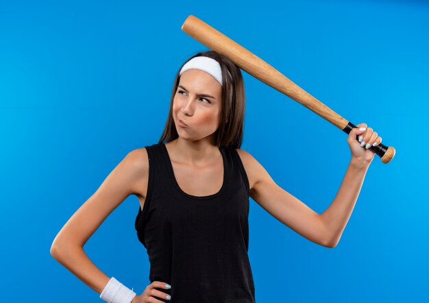 Confused young pretty sporty girl wearing headband and wristband holding baseball bat and touching her head with it looking at side with hand on waist isolated on blue wall