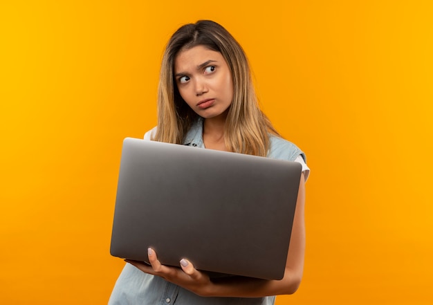 Смущенная молодая симпатичная студентка в задней сумке, держащая ноутбук, смотрит в сторону, изолированную на оранжевом, с копией пространства