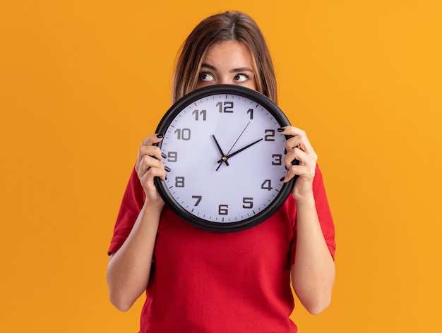 Confused young pretty woman holds clock looking at side isolated on orange wall