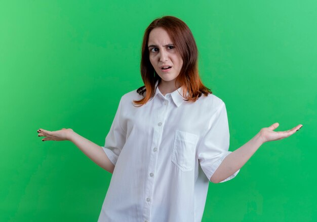 Confused young redhead girl spreads hands isolated on green background