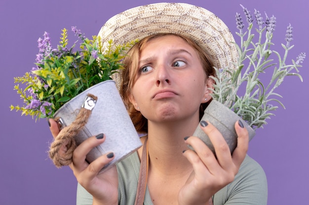 紫色の植木鉢に花を保持しているガーデニング帽子をかぶっている混乱した若いスラブ女性の庭師