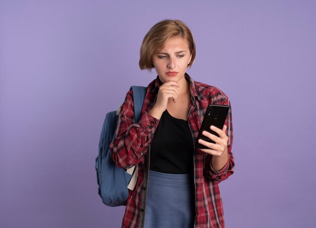 Смущенная молодая славянская студентка в рюкзаке кладет руку на подбородок и смотрит на телефон