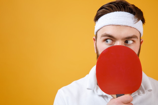 Confused young sportsman holding racket for table tennis covering mouth