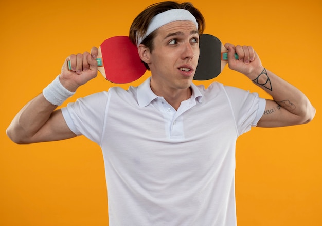 Foto gratuita confuso giovane ragazzo sportivo guardando lato indossando la fascia e il braccialetto che tengono le racchette da ping pong sul collo