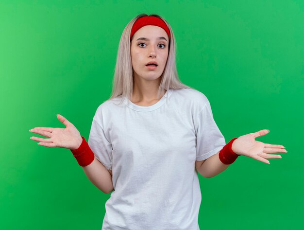 Confused young sporty woman with braces wearing headband and wristbands holds hands open isolated on green wall