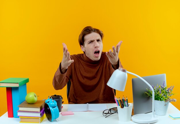 Confused young student boy sitting at desk with school tools holding out hands