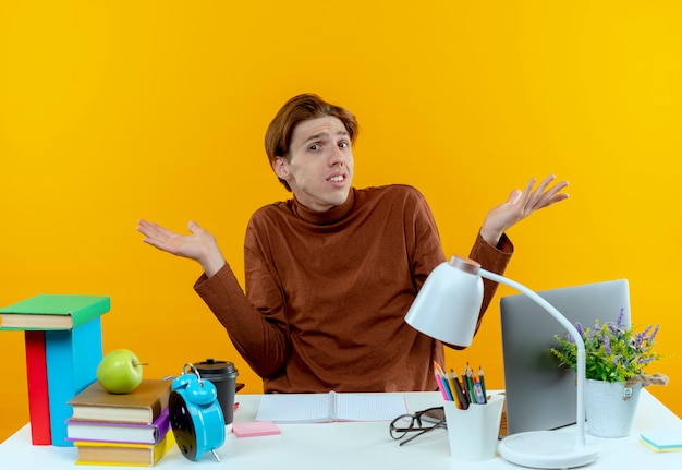 confused young student boy sitting at desk with school tools spreads hands