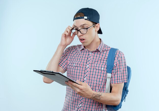 Confused young student boy wearing backpack and glasses and cap holding and looking at clipboard 