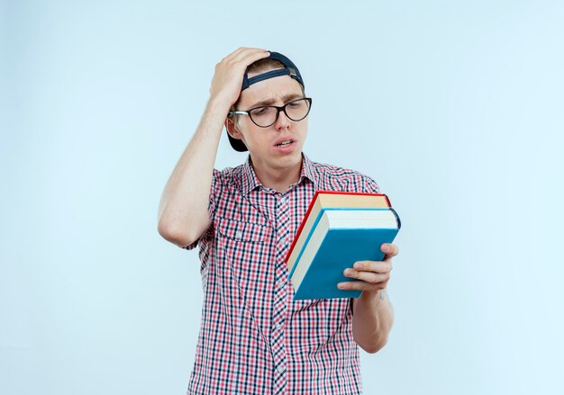 Confused young student boy wearing glasses and cap holding and looking at books