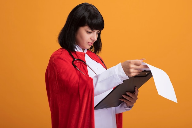 Confused young superhero girl wearing stethoscope with medical robe and cloak flipping through clipboard