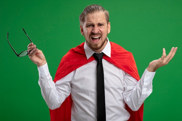 Free photo confused young superhero guy wearing tie holding glasses and spreading hand isolated on green