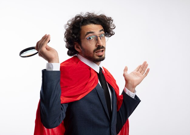 Confused young superhero man in optical glasses wearing suit with red cloak holds magnifying glass and keeps hand open isolated on white wall