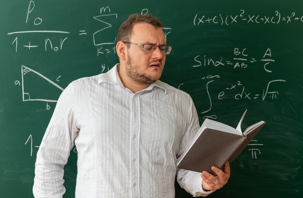 confused young teacher wearing glasses standing in front of chalkboard in classroom reading notepad