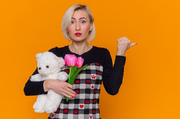 Confused young woman in beautiful dress holding bouquet of tulips and teddy bear as gifts pointing back with thumb celebrating international women's day standing over orange wall