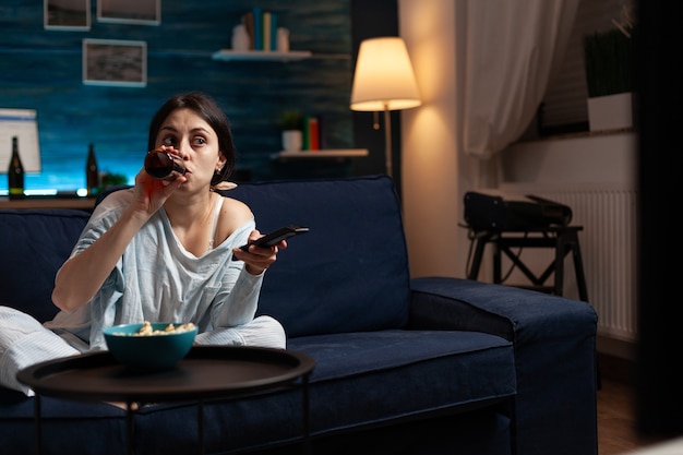 Confused young woman drinking beer while watching movie on tv