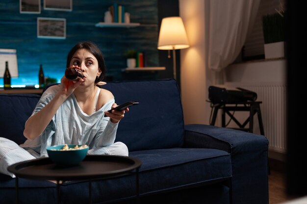 Confused young woman drinking beer while watching movie on tv