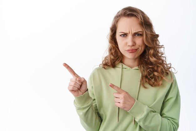 Confused young woman frowning, judging, smirk and look upset and displeased, pointing at upper left corner promotional text, accuse someone, white wall