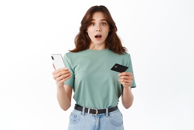 Смущенная молодая женщина держит пластиковую кредитную карту и смартфон с открытым ртом и смотрит с нерешительным взволнованным лицом на камеру, стоящую на белом фоне