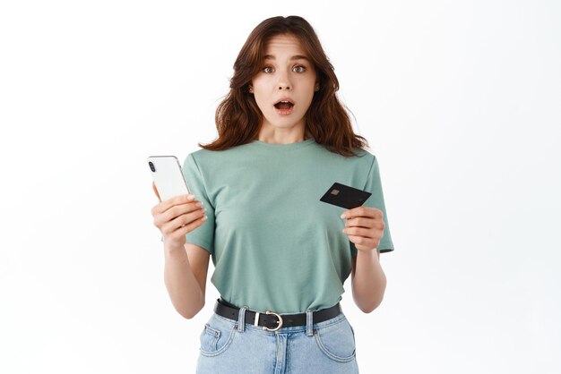 Confused young woman holding plastic credit card and smartphone open mouth and looking with indecisive worried face at camera standing against white background