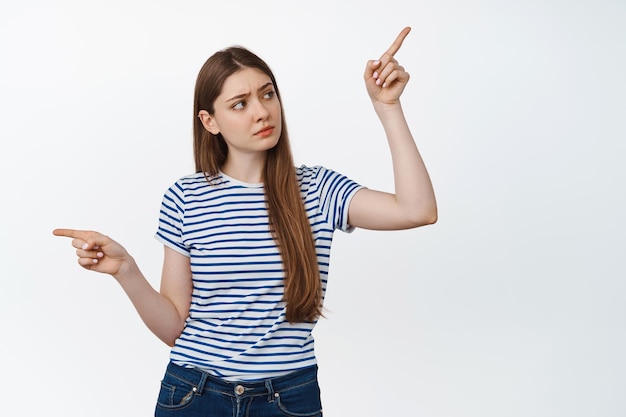 Confused young woman making decision, pointing sideways, looking up thoughtful, choosing, standing over white background.