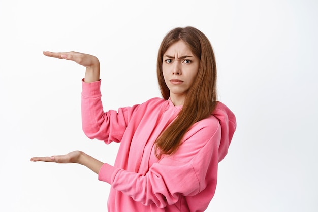 Confused young woman showing object empty box place for copyspace look puzzled at camera holding product on display standing against white background