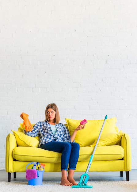 Confused young woman sitting on yellow sofa holding gloves and brush