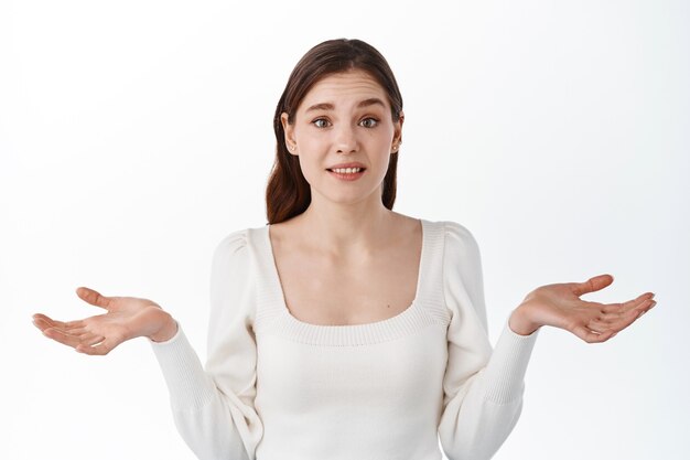 Confused young woman student shrugging shoulders, not know what to do, standing clueless and puzzled against white wall