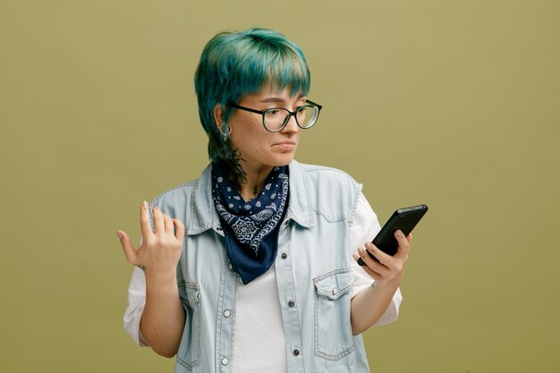 Смущенная молодая женщина в очках, бандане на шее, держащая руку в воздухе с помощью мобильного телефона, изолированного на оливково-зеленом фоне