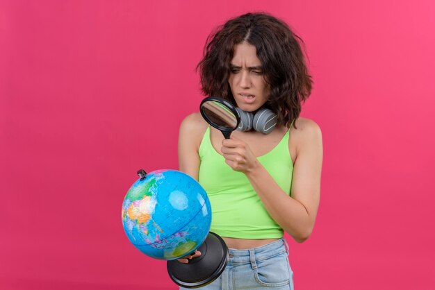 A confused young woman with short hair in green crop top in headphones looking at globe with magnifying glass