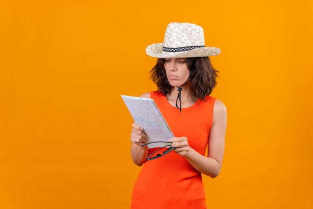 A confused young woman with short hair in an orange shirt wearing sun hat holding sunglasses looking at map 