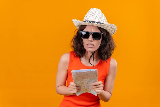 A confused young woman with short hair in an orange shirt wearing sun hat and sunglasses holding map 