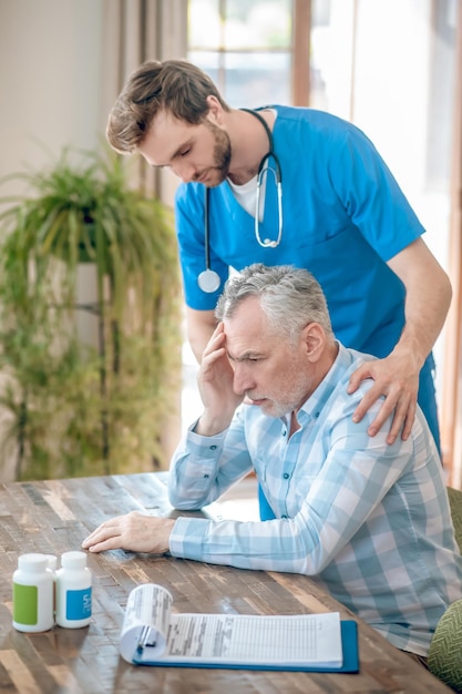 Considerate dark-haired bearded young Caucasian geriatrician with a stethoscope leaning over a sad mature patient