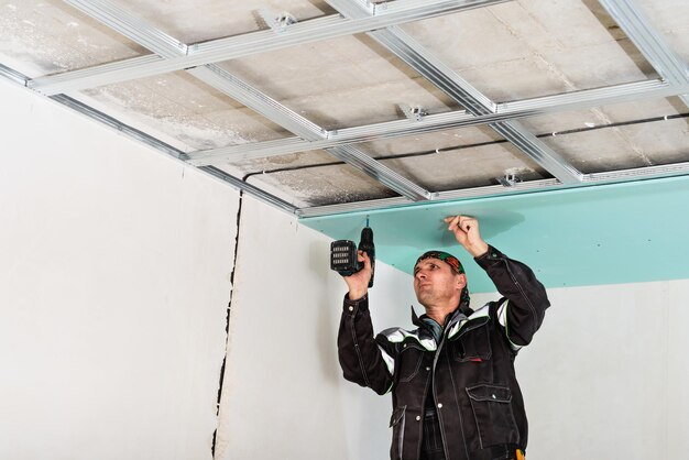 建設作業員のアセンブリは、乾式壁で天井を吊り下げ、ドライバーで乾式壁を天井の金属フレームに固定します。