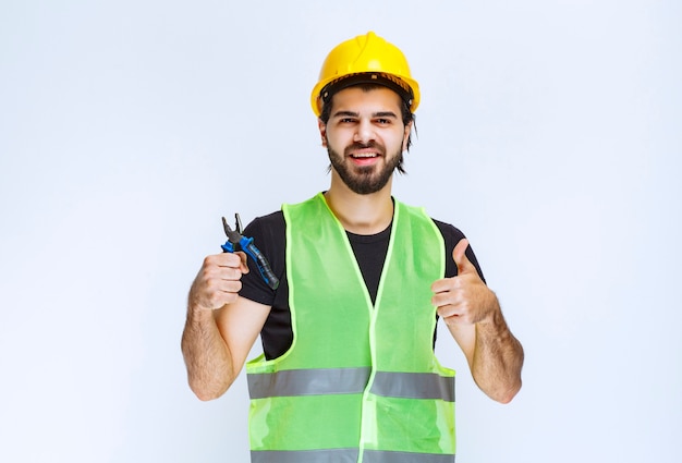 Рабочий-строитель держа голубые плоскогубцы и показывая большой палец вверх.