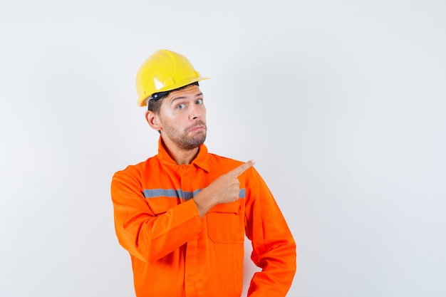 Construction worker pointing away in uniform, helmet and looking hesitant , front view.